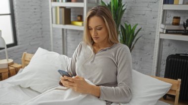 Kadın rahat bir yatak odasında, kitap ve bitkilerle çevrili, rahat bir yatak odasında akıllı telefon kullanarak rahatlıyor..