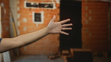 İnşaat alanında kısmen inşa edilmiş bir arka planla adamın eli içeri uzanıyor. Çalışma ortamında faaliyet ya da iletişim kurulduğunu gösteriyor..