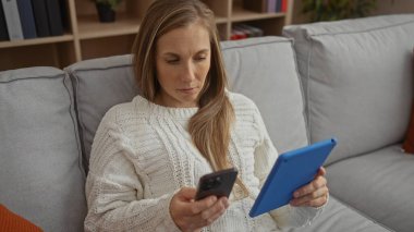 Oturma odası ortamında telefonla tablet okuyan, rahat giyimli ve rahat atmosferli bir kadın modern iç mekanda teknolojik alet kullanımını gösteriyor..