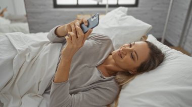 Kadın rahat yatak odasında akıllı telefon kullanıyor. Parlak doğal ışığı var. Evde rahat ve rahat bir ortam var..