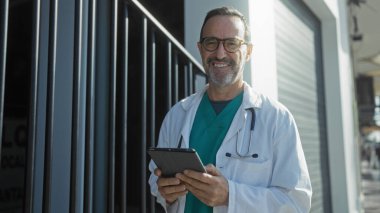 Dışarıda elinde tabletle bekleyen doktor şehir ortamında gülümsüyor beyaz önlüklü ve steteskoplu olgun bir erkek hastane binasının dışında tıbbi bir meslek olduğunu gösteriyor..