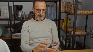 Ofisteki olgun İspanyol adam, kavanoz ve mutfak gereçleriyle dolu rafların arasında akıllı telefona odaklanıyor..