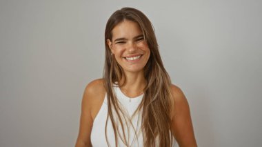 Genç İspanyol kadın gülümsüyor, beyaz arka plana karşı duruyor, beyaz kolsuz bluz giyiyor, omuzlarında kumral saçlar, rahat bir ortamda kendine güveni ve güzelliği yayıyor.