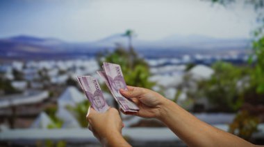 Arka planda şehir manzarası bulanıklaşan Endonezya rupiasını sayan eller, para değişimi veya sayım içeren kentsel iş veya mali işlemleri akla getiriyor.