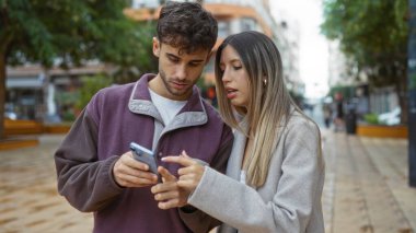 Şehir caddesinde akıllı telefonu birlikte kontrol eden erkek ve kadın, modern ilişki ve iletişimi açık şehir ortamında günlük kıyafetlerle ve rahat atmosferle sergiliyor..