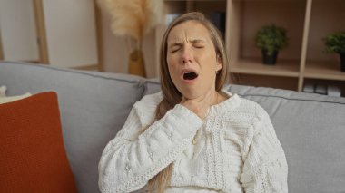 Bir kadın evdeki kanepede otururken boğazında ağrı hissediyor. Rahatsızlık ve hastalık gösteriyor. Sıcak bir oturma odasında, sıcak bir süveter giyiyor..
