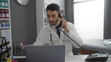 Hastane odasında dizüstü bilgisayarla çalışan, beyaz önlüklü ve telefonda konuşan, tıbbi ekipman ve ofis malzemeleriyle çevrili genç bir adam..