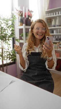 Dekor dükkanında elinde telefonla gülümseyen genç bir kadın, gözlük ve önlük takıyor, ev bitkileri ve dekorasyon malzemeleriyle çevrili.