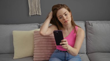 Evdeki koltukta oturan kızıl saçlı genç bir kadın, cep telefonuna bakarken saçlarını eliyle tarıyor..