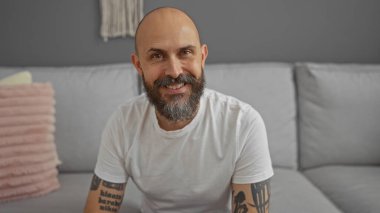 Yakışıklı, kel, sakallı ve dövmeli adam oturma odasında oturmuş gülümsüyor.