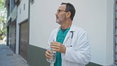 Steteskoplu beyaz önlüklü bir adam şehir sokağında elinde su şişesi tutarak düşünceli bir şekilde duruyor..