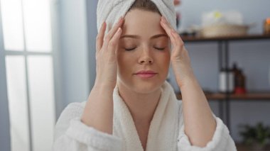 Kafasına havlu geçirmiş genç bir kadın, arka planda sağlık ürünleri olan bir spa odasında rahatlatıcı bir anın tadını çıkarıyor.