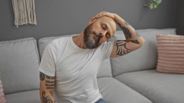 Yakışıklı, kel, sakallı ve dövmeli bir adam evin içinde bir kanepede otururken apartman dairesinde esniyordu.