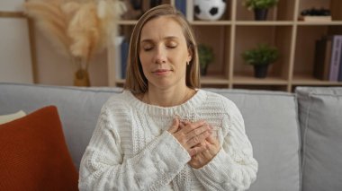 Oturma odasının içinde elleri ve gözleri kapalı meditasyon yapan, huzur ve rahatlık saçan, modern ev dekoru ve yumuşak ışıklandırmayla çevrili bir kadın..