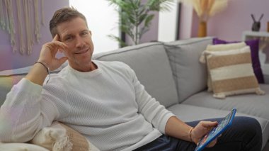 Yakışıklı adam modern oturma odasında rahat bir yaşam tarzını yansıtan şık dekorlu tablet tutuyor.