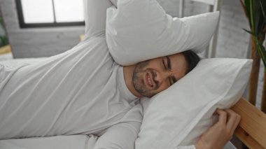 Yatak odasındaki genç adam beyaz gömlekli, kafasında yastıkla yatıyordu. Gürültüyle ya da modern dekorlu bir iç mekanda rahatsız bir şekilde uyanmış gibi görünüyordu.