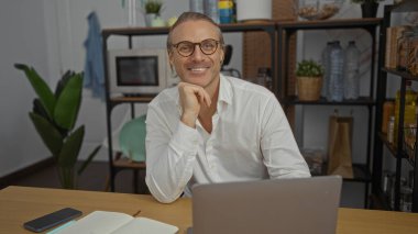 Modern bir ofiste masasında oturan olgun bir adam. Etrafı bitkilerle, kitaplarla ve bilgisayarlarla çevrili sarı saçları ve gözlükleriyle güvenle gülümsüyor..