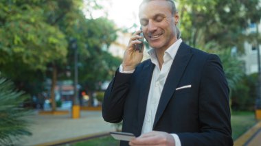 Şehir parkında sarışın, orta yaşlı bir adam kredi kartına bakarken telefonla konuşuyor. Takım elbiseli, şehir geçmişi parlak gün ışığında bulanık..