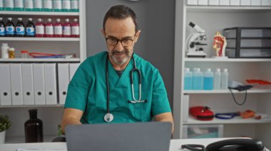 Tıbbi ekipman ve malzeme ile çevrili hastane odasında dizüstü bilgisayarda çalışan yetişkin bir İspanyol erkek doktor profesyonel sağlık ortamını ön plana çıkarıyor.