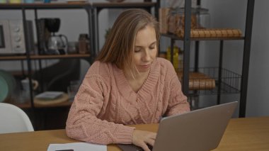 Ofis ortamında çalışan bir kadın, dizüstü bilgisayarı kullanarak etrafı raflarla ve ofis malzemeleriyle çevrili, düşünceli bir ifadeyle iç mekanda çalışıyor.