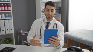 Klinikteki genç İspanyol adam, modern sağlık teknolojisi ve hasta iletişimini vurgulayan profesyonel bir ortamda video danışmanlığı için tablet kullanıyor..
