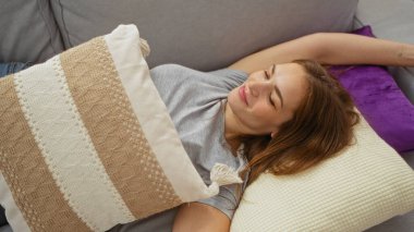 Kadın, sakin bir şekilde gülümserken ve evin içinde huzurlu bir atmosferin keyfini çıkarırken, iç dekoratif yastıklarla parlak bir dairede koltukta dinleniyor..