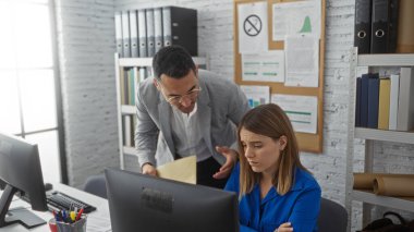 Takım elbiseli bir adam bilgisayarda oturan kadınla konuşuyor. Duvarında belgeler ve çizelgeler var..