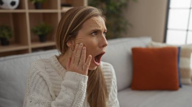 Oturma odasında oturan kadın şok olmuş görünüyor, el üstünde, lüks bir dairede şık bir dekorasyonla şaşkınlığını ifade ediyor, parlak gün ışığı alan bir yer..