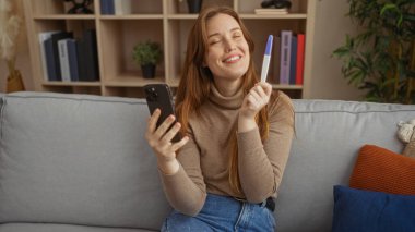 Sıcak bir oturma odasındaki kanepede oturan mutlu bir kadın, pozitif bir hamilelik testi ve akıllı bir telefon tutuyor, sıcak ve davetkar bir iç mekanla çevrili, neşe ve şaşkınlığı ifade ediyor..