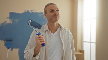 Orta yaşlı bir adam mavi boya silindiriyle yeni bir ev boyuyor, oturma odasında dikiliyor, günlük beyaz gömlek giyiyor ve duvarlardaki gelişime hoşnutça bakıyor..
