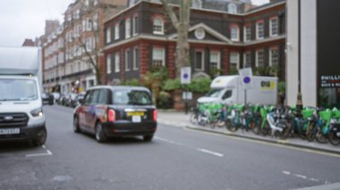 Güz mevsiminde bir Londra caddesinin bulanık görüntüsü ikonik siyah taksi, park edilmiş bisikletler ve gri gökyüzünün altında tarihi mimari.