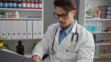 Klinik odasında çalışan, steteskop ve beyaz önlük giyen, tıbbi malzeme ve kitaplarla çevrili, kapalı alanda profesyonel bir ortamda çalışan genç bir İspanyol.