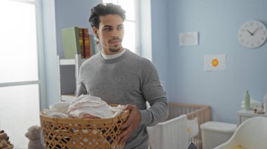 Çamaşır odasında elinde sepet tutan genç İspanyol bir adam, evde, iyi aydınlatılmış, temiz bir ortamda ev işlerini öneriyor.