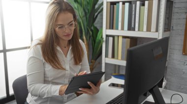 Modern bir ofis ortamında çalışan, masasında tablet kullanan, kitaplarla çevrili, kapalı bir iş ortamında odak ve profesyonellik gösteren bir kadın..