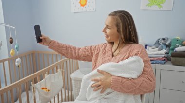Kadın, bebeğiyle rahat bir yatak odasında selfie çekerken gülümsüyor, arka planda beşiği ve oyuncakları olan değerli anı yakalıyor, annelik sevincini sergiliyor..