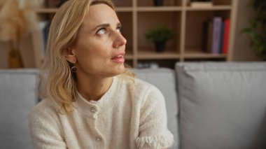 İçerde oturan bir kadın, rahat bir oturma odasında, sarı saçlı ve açık renk süveterli bir kadın, rahat bir ev ortamında güzel, olgun bir anı yakalıyor..