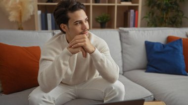 Beyaz bir kazak giyen, düşünceli ve rahat görünen, rahat bir oturma odasında renkli yastıklarla bir kanepede oturan İspanyol genç adam..