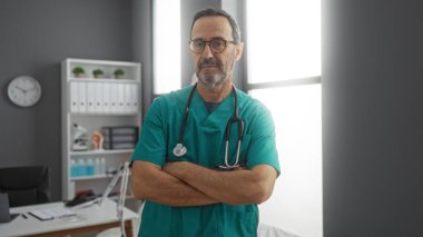 İspanyol erkek doktor güvenli bir şekilde kollarını kavuşturarak aydınlık bir hastane odasında duruyor, tıbbî profesyonelliğin ve kapalı alanda uzmanlığın özünü somutlaştırıyor..