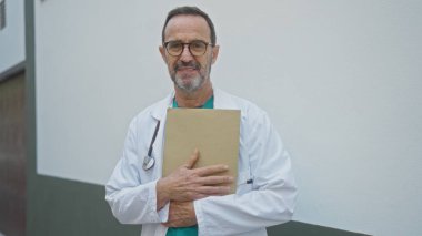 Beyaz önlüklü yaşlı İspanyol erkek doktor elinde bir dosya klasörü, somut olgunluk ve profesyonelliği taşıyan bir şehir caddesinde güvenle duruyor..