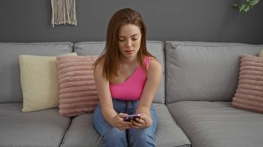Genç bir kadın oturma odasında pembe bir bluz ve kot pantolon giyiyor, cep telefonu kullanıyor, odaklanmış ve düşünceli görünüyor, arka planda gri bir kanepe ve dekoratif yastıklar var..