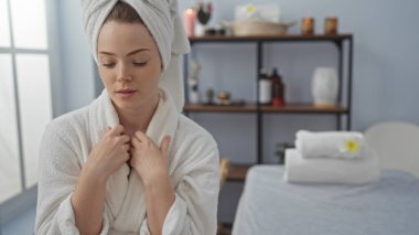 Kaplıcada kafasında bornoz ve havlu olan genç bir kadın, arka planda raflar ve havlularla bir sağlık merkezinde rahat görünüyor.