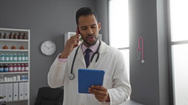 Klinikteki İspanyol doktor, tıbbi malzemeler ve modern iç mekânlarla çevrili steteskopla tableti kontrol ederken telefonla konuşuyor ve çalışmalara odaklanıyor.