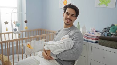 Genç adam bebeği yatak odasında tutuyor, sıcak bir aile sahnesi sergiliyor sıcak bir iç mekan ve yumuşak ışıklandırmayla, ebeveynler ve çocuklar arasındaki bağı vurguluyor..