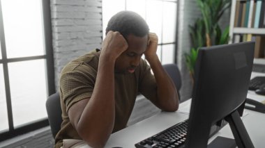 Modern ofiste masa başında oturan genç adam bilgisayar karşısında hayal kırıklığına uğramış ve elleri başının üstünde duruyor.