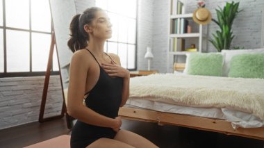 Huzurlu bir yatak odasında, yatağın yanında meditasyon yapan bir kadın. Parlak doğal ışık ve modern ev dekorasyonu unsurlarıyla sakin bir ortam sergiliyor..