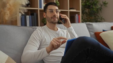 Oturma odasındaki bir koltukta oturan genç İspanyol bir adam telefonla konuşuyor ve tablet kullanıyor. Kitaplar ve dekorlarla modern bir ev dekorasyonu sergiliyor..