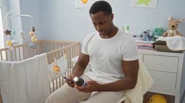 Yakışıklı bir adam rahat bir yatak odasında oturuyor. Yakınlarda bir bebek beşiği ile telefonunu kontrol ediyor. Güzel dekore edilmiş ve evi hoş karşılıyor..