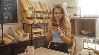 Fırındaki sarışın kadın, ahşap bir masada otururken akıllı telefon kullanıyor. Raflarda çeşitli ekmek ve hamur işleri sergileniyor.