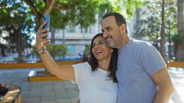 İspanyol çift, şehir parkında selfie çekerek aşk ve birlikteliğin tadını çıkarıyor. Açık hava ortamındaki bağlantılarını ve neşelerini vurguluyorlar..