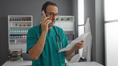 Klinikte önlük giyen İspanyol bir adam telefonla konuşurken rafları ve doğal ışığı olan ve profesyonelliği somutlaştıran parlak, organize tıp odasındaki belgeleri inceliyor..